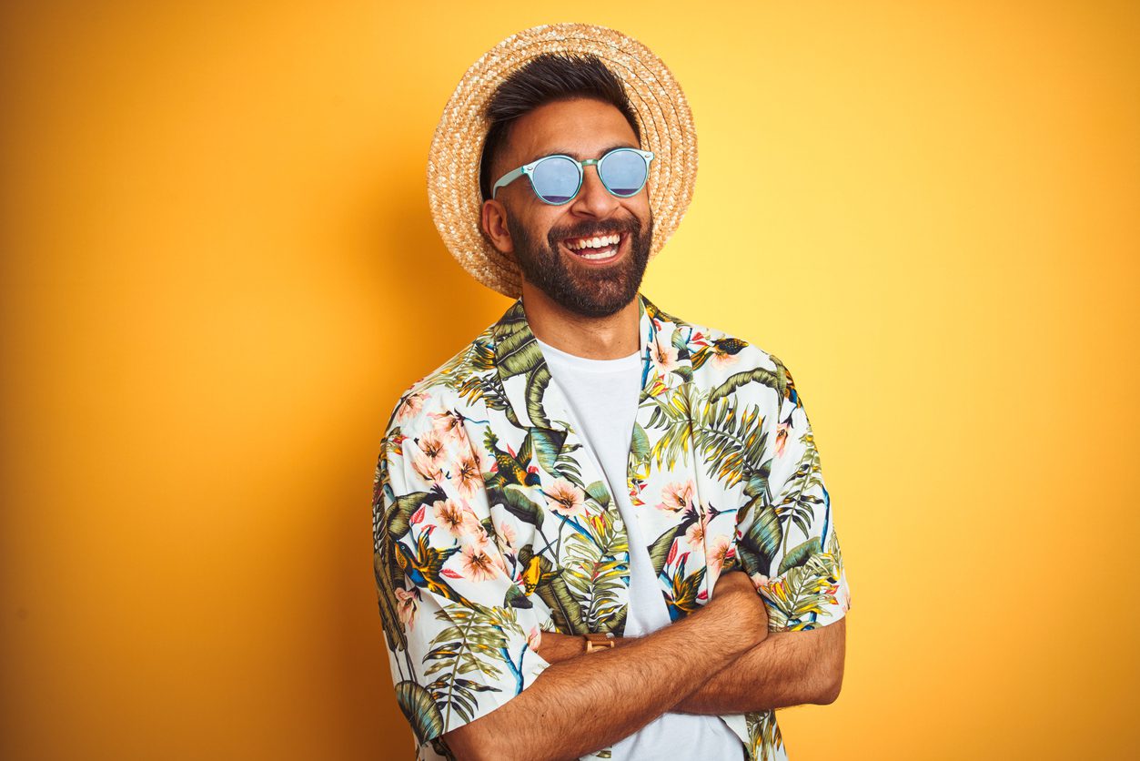 An Indian man dressed in summer clothing such as a floral shirt,hat,and sunglasses poses against a yellow background.
