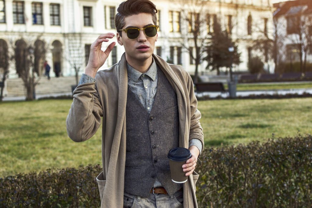 Young stylish man wearing sunglasses and academia-inspired clothing, holding a coffee in the city.
