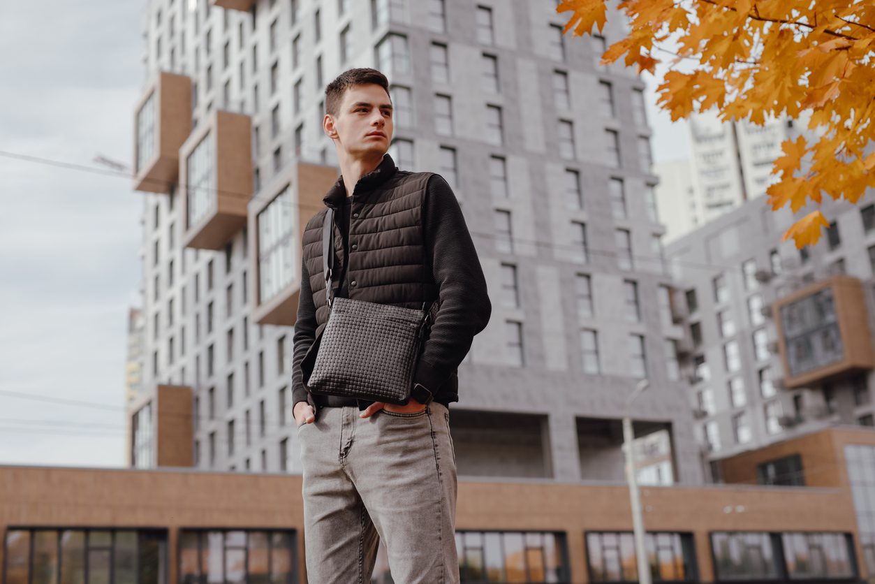 Young man wearing a quilted vest while holding a black messenger bag outdoors.